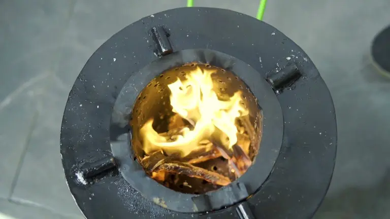 Top-down view of a biomass gasifier stove project showing active yellow flames burning wood fuel in a black metal chamber.