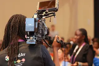 A videographer with dreadlocks filming a speaker at a corporate event using a professional cinema camera and monitor.